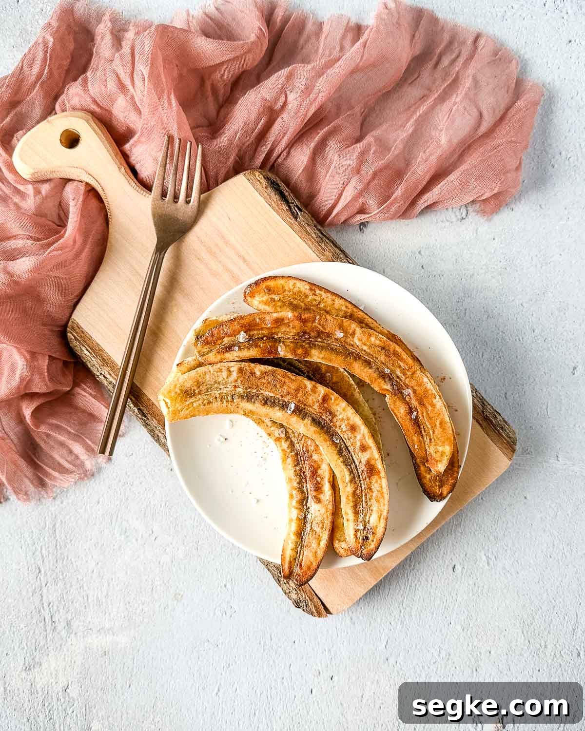 Air fryer bananas on a white plate over a rustic wooden cutting board with a pink linen and a copper fork.