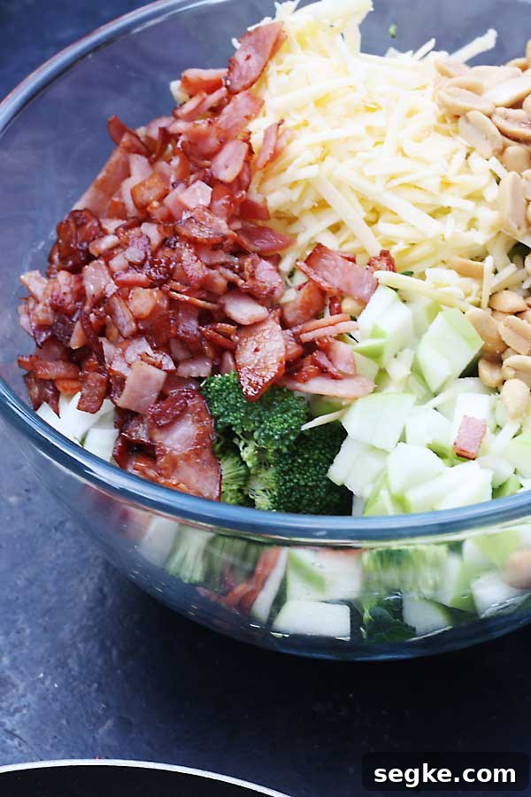 All the ingredients needed to make a broccoli, apple and bacon salad in a glass bowl on a black background.