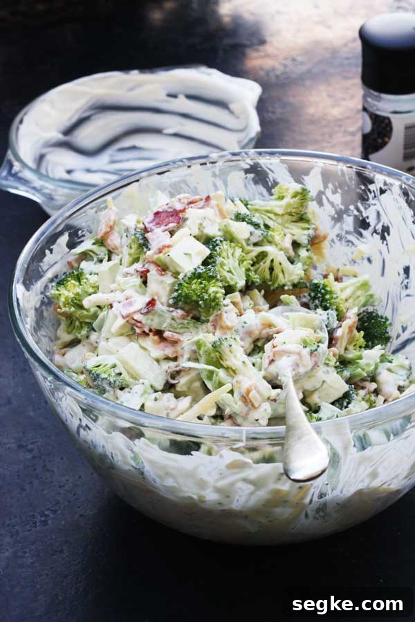 Broccoli and bacon and apple salad in a glass bowl with a big metal spoon in it.