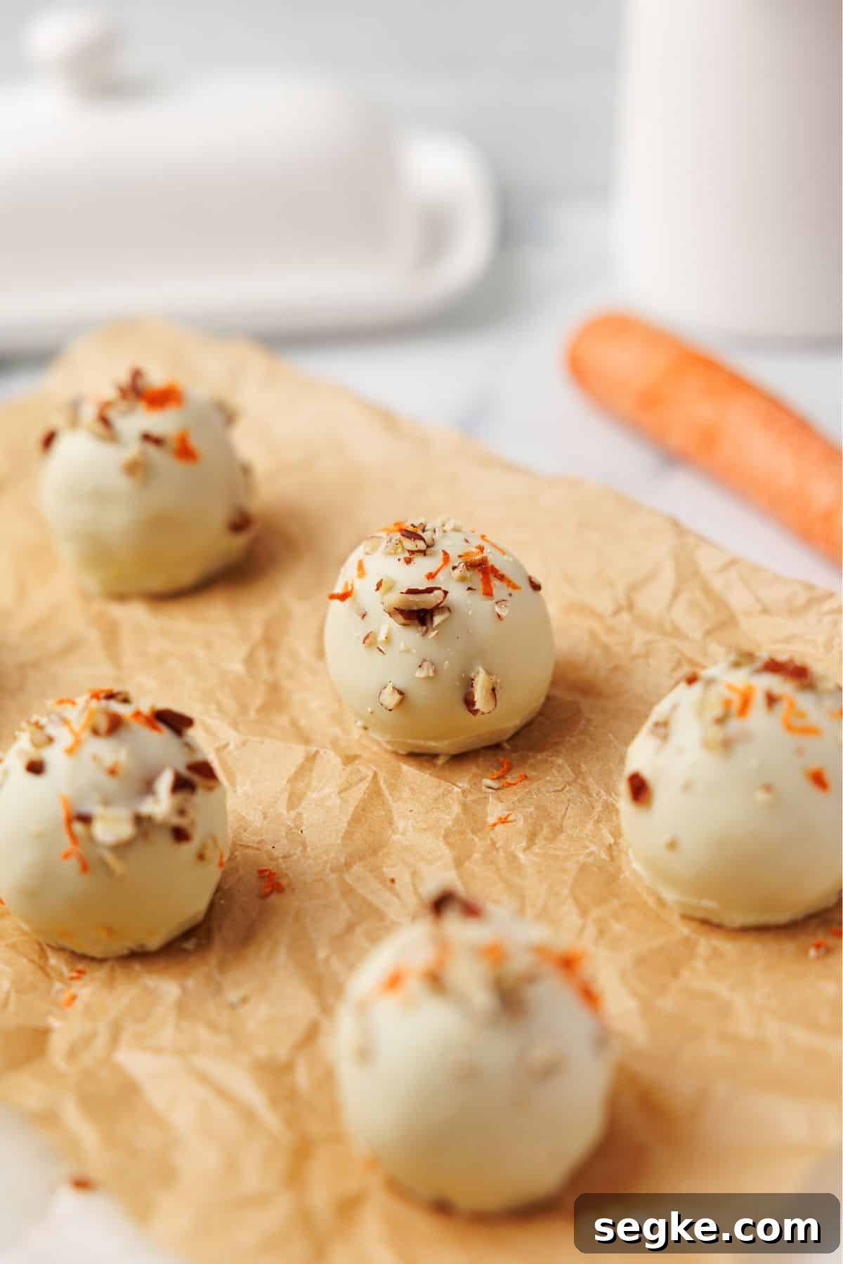 No Bake Keto Carrot Cake Protein Bites 8 Six white chocolate carrot cake bites on parchment paper topped with pecans and shredded carrot, with a whole carrot in the background