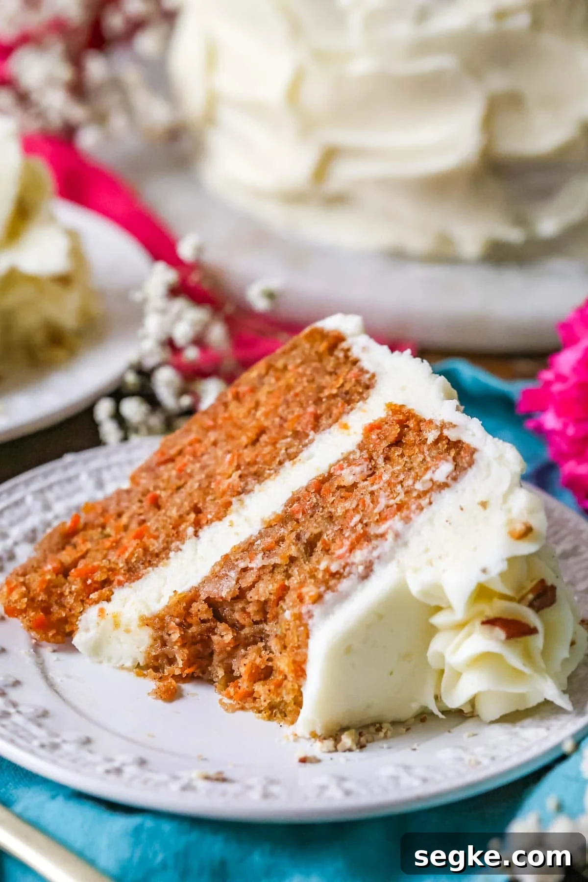 This is an incredibly moist, perfect, BEST Carrot Cake recipe, hands down! A slice of the best carrot cake recipe on a white plate with white flowers in the background. A bite missing from the slice.