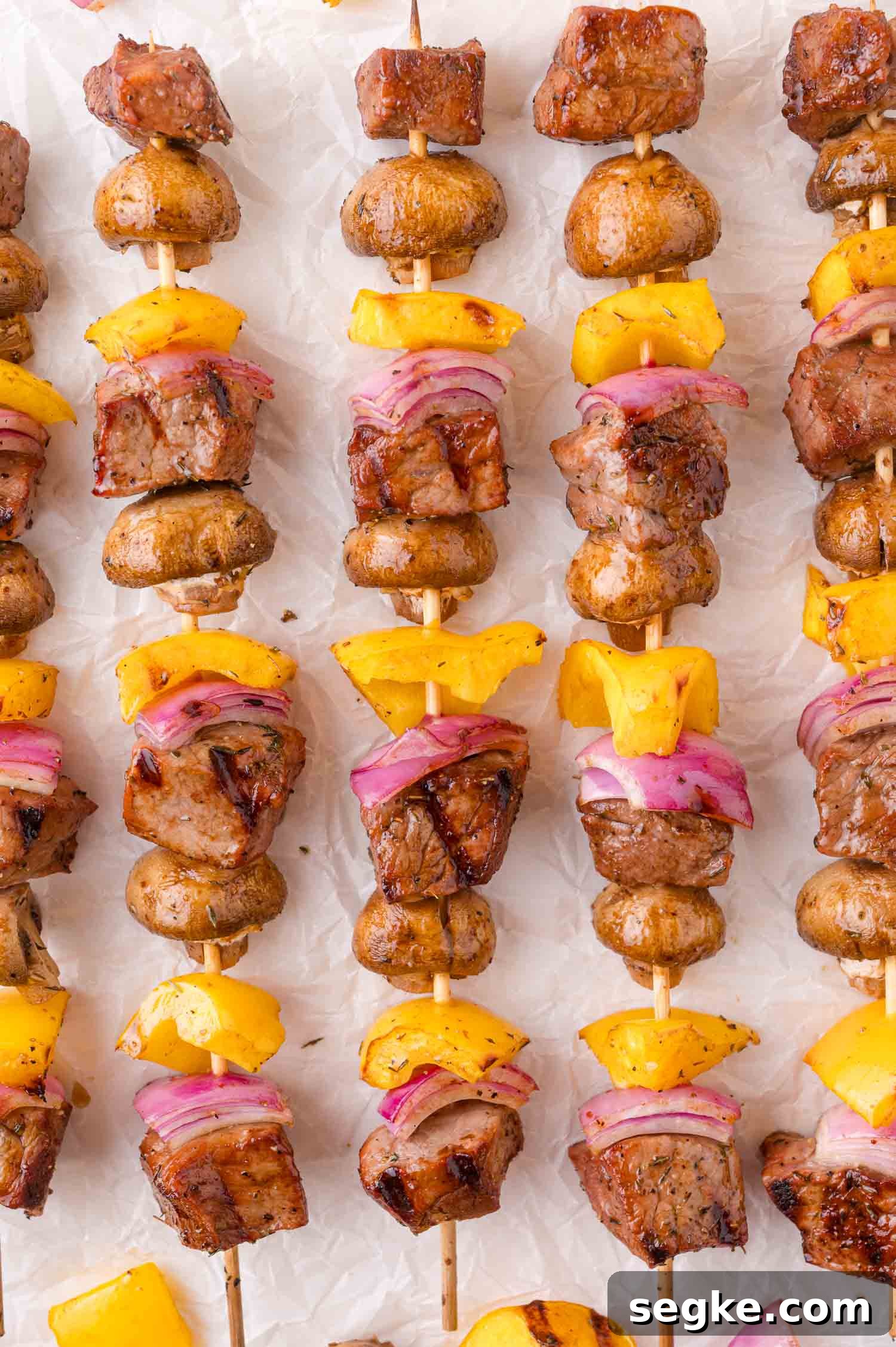 Marinated Steak Kabobs with Garlic and Herbs 7 Skewers with grilled beef, mushrooms, yellow bell peppers, and red onions arranged in rows on a white surface.