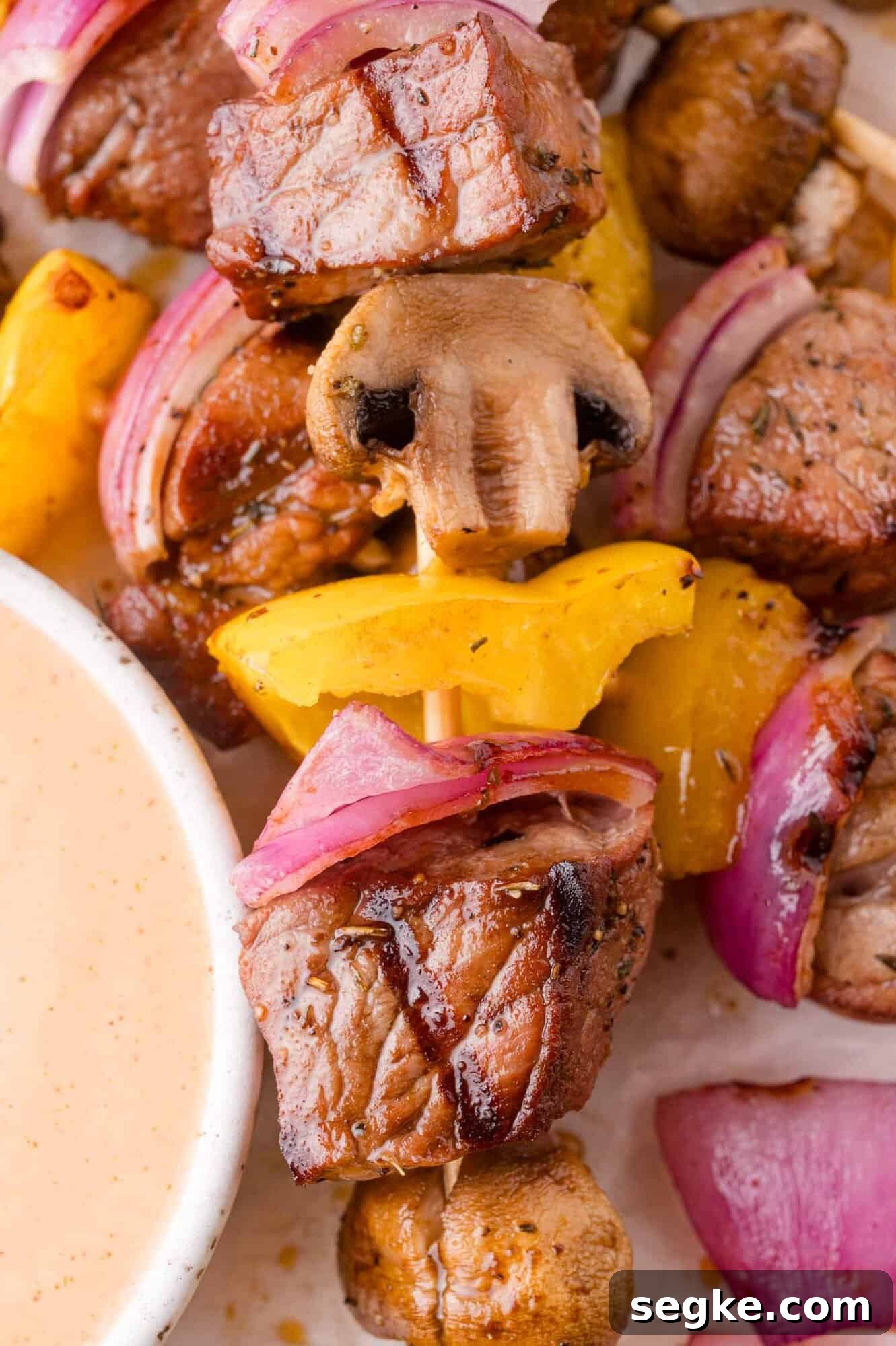 Marinated Steak Kabobs with Garlic and Herbs 3 Close-up of a skewer with grilled beef, yellow bell pepper, red onion, and mushroom next to a bowl of dipping sauce.