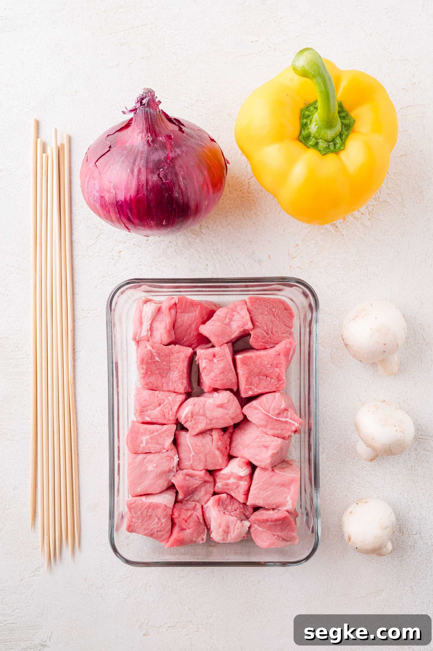 Marinated Steak Kabobs with Garlic and Herbs 4 Glass container with raw steak cubes, a red onion, yellow bell pepper, two white mushrooms, and wooden skewers arranged on a white surface.