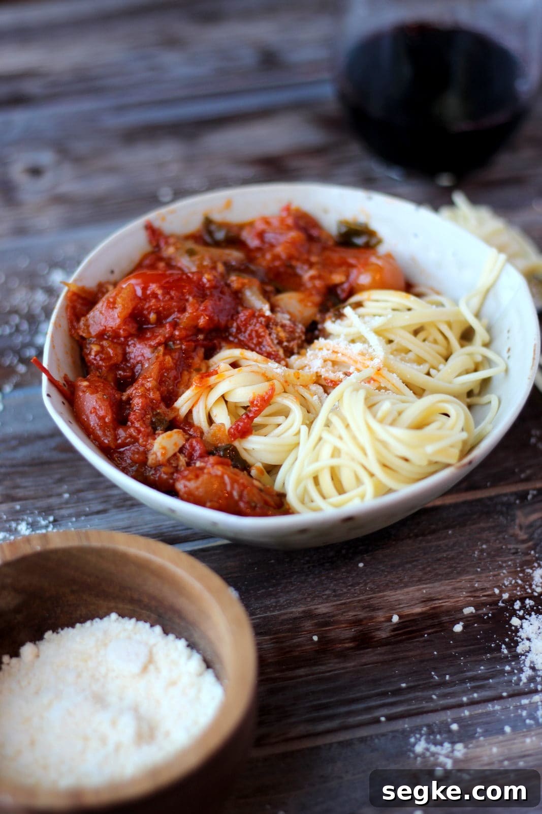 A bowl of cooked spaghetti noodles with roasted tomato sauce next to a dish of grated parmesan cheese.