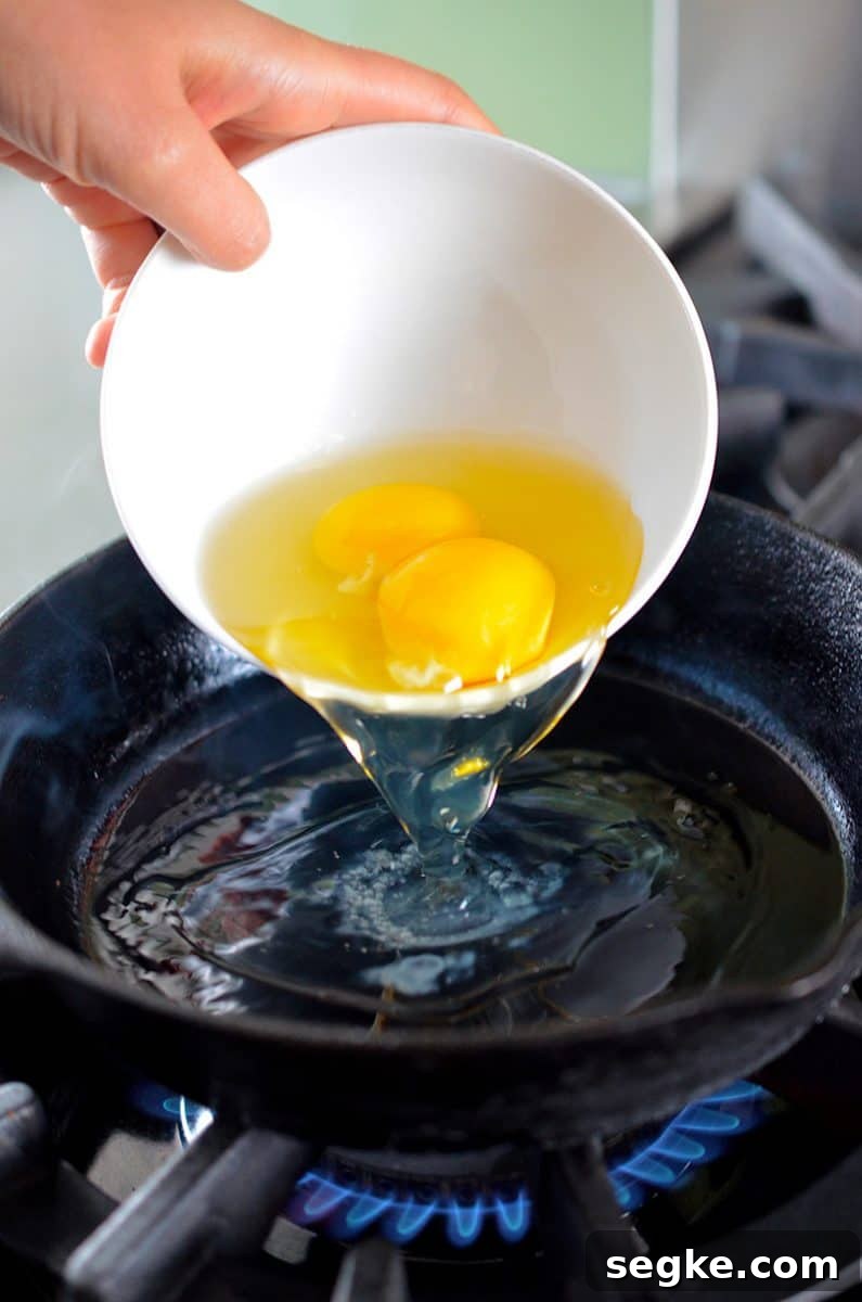 Two eggs in a white bowl are poured into a hot cast iron skillet.