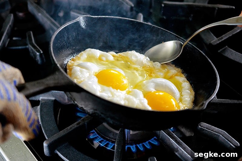 A cast iron skillet filled with two sunnyside up fried eggs is tilted with a spoon ladling hot ghee on the uncooked whites.