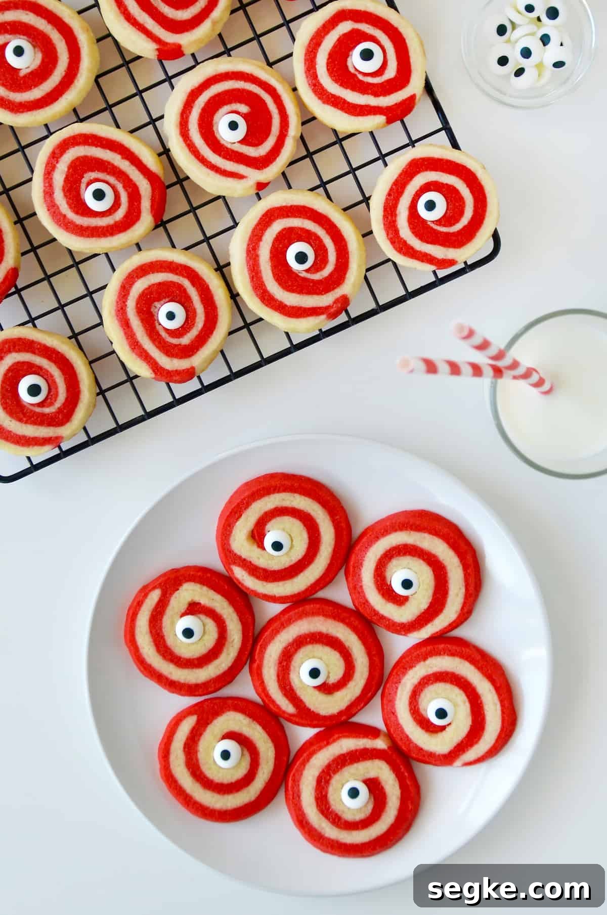 Spooky Halloween Dessert Recipes to Impress Guests 11 A plate of red and white Halloween pinwheel cookies with monster eyes and a glass of milk and a cooling rack with more cookies on it above it.