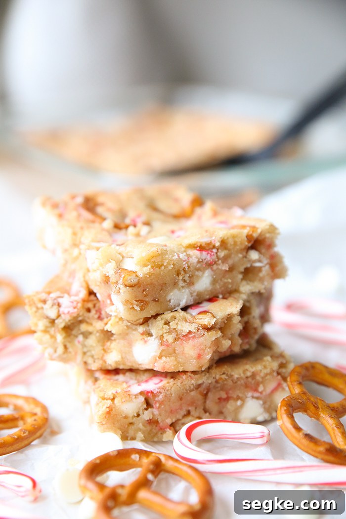Three peppermint white chocolate pretzel cookie bars stacked on each other with small candy canes and pretzel twists surrounding them. Faded in the background is a glass 9x13 baking dish of the cookie bars and a black serving spatula.