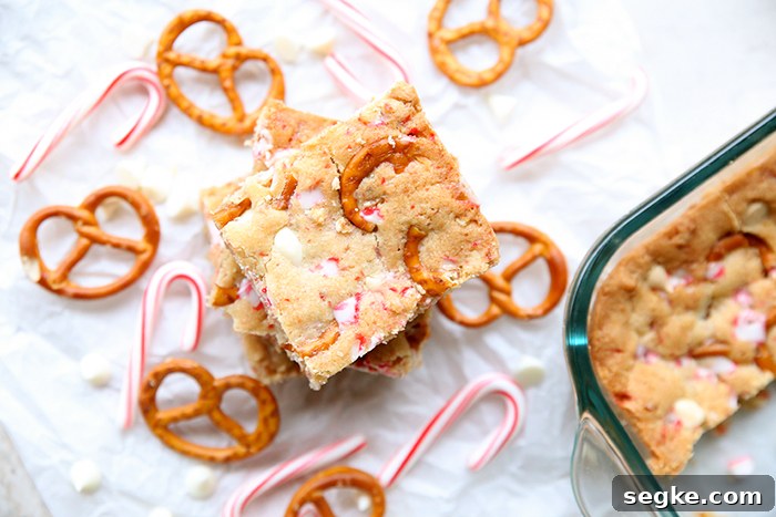 Pretzel twists and small candy canes surrounding an offset stack of white chocolate peppermint pretzel bars with the corner of a glass 9x13 baking dish of the bars in the right-hand corner.