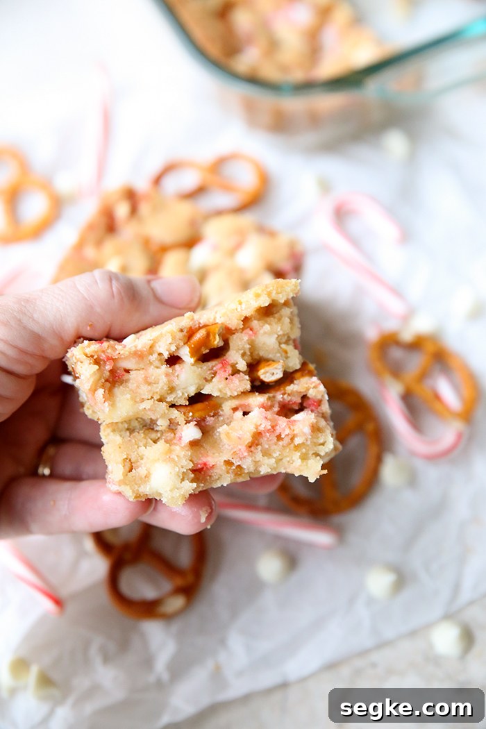 A broken in half white chocolate candy cane pretzel cookie bar held above scattered white chocolate chips, pretzel twists and small candy canes. In the upper right corner is a glass 9x13 dish corner with the cookie bar.