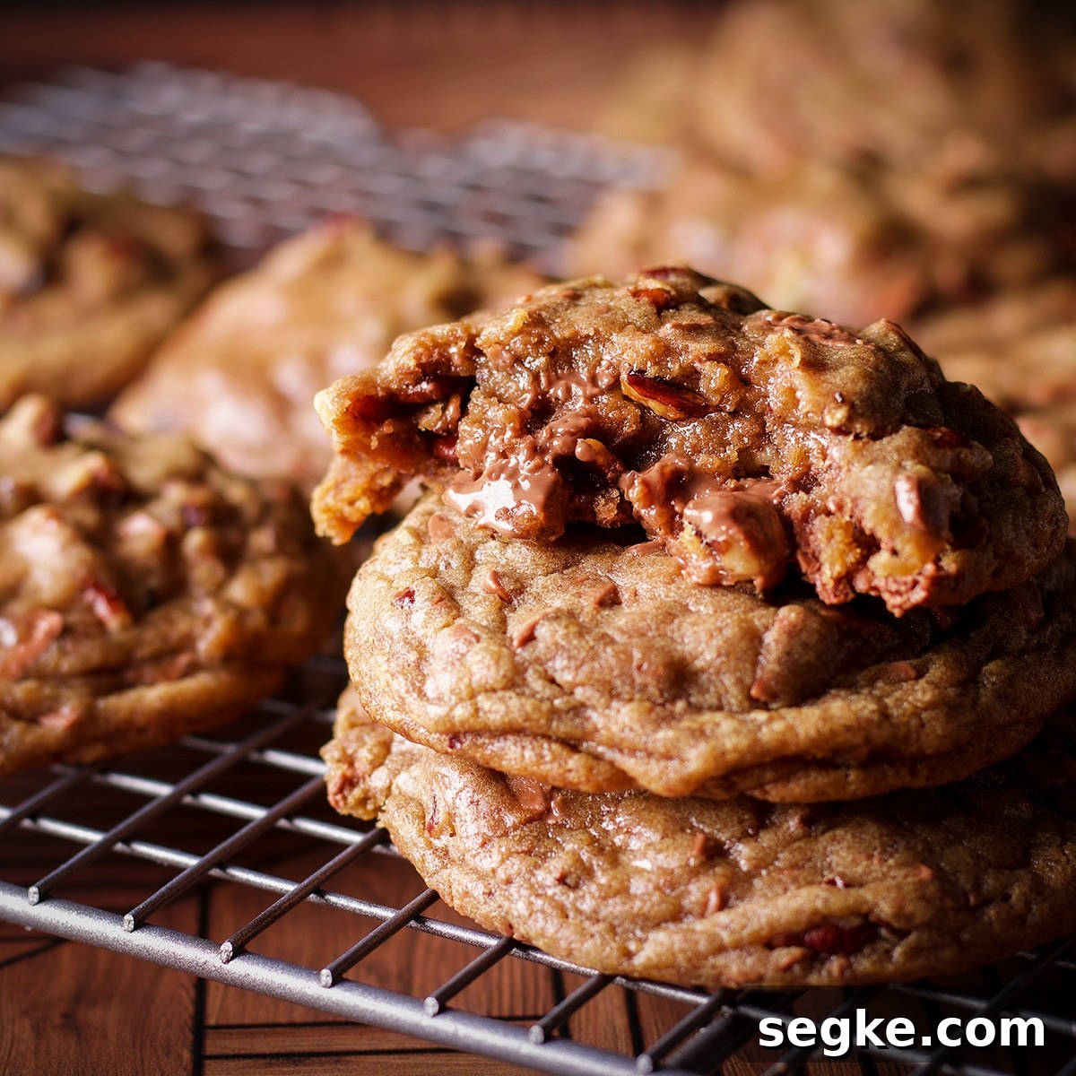 A wire rack with freshly baked chocolate chip pecan cookies cooling on it. One of the cookies has been broken in half so you can see the gooey center.