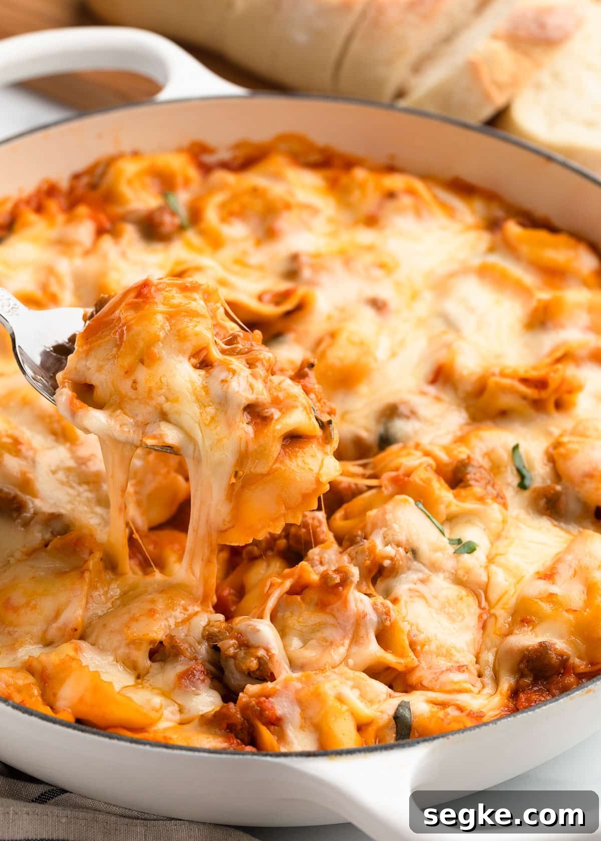a spoonful of baked tortellini with sausage being scooped from a white enameled cast iron pan with a cheese pull.