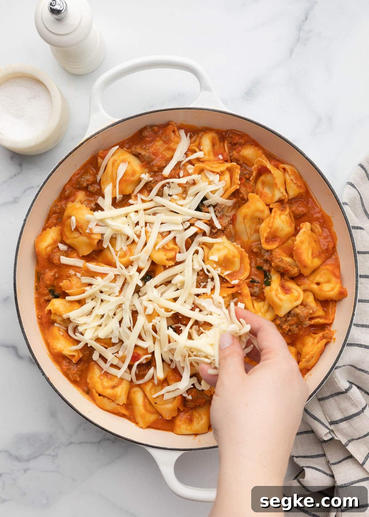 sprinkling mozzarella over sauced tortellini in a white enameled cast iron pan