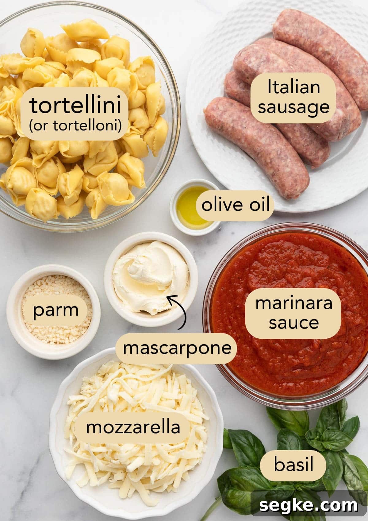 overhead of ingredients needed to make baked tortellini with sausage in bowls on a white marble board. Ingredients have text label overlays.