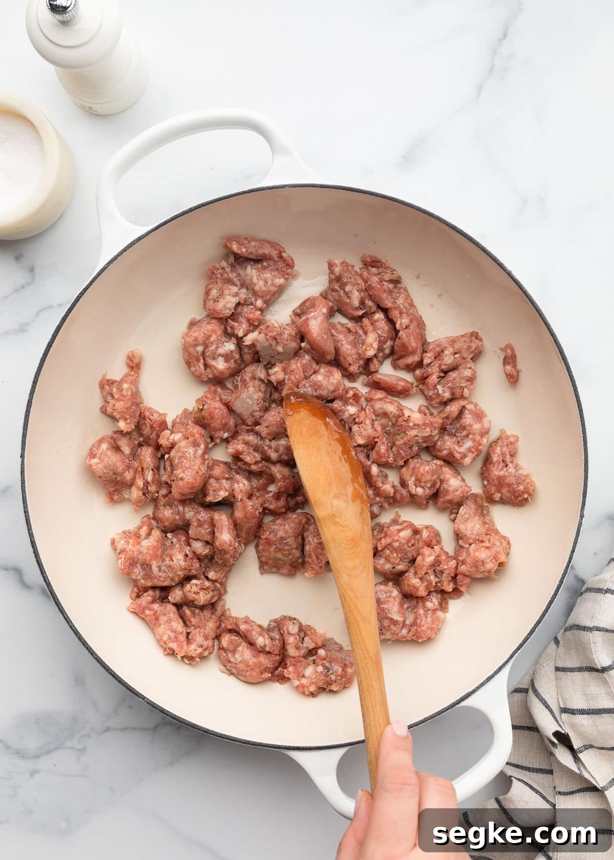 raw Italian sausage in a white enameled cast iron pan being stirred with a wooden spoon