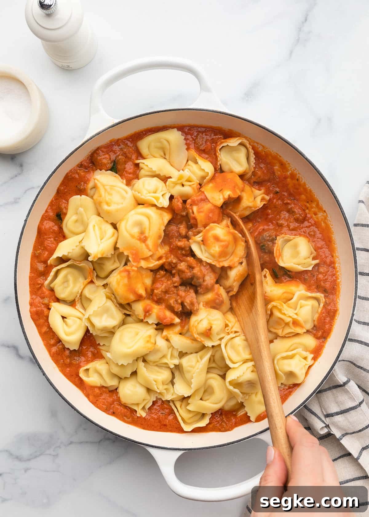 stirring cooked tortellini into sausage marinara sauce with a wooden spoon in a white enameled cast iron pan