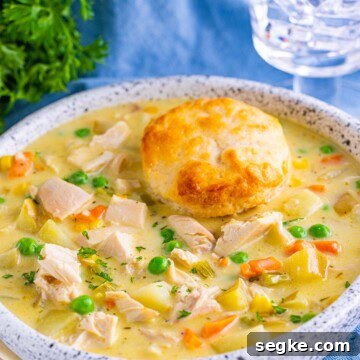 A white bowl of chicken pot pie soup topped with a flaky biscuit, with peas, corn, and shredded chicken in a creamy broth.