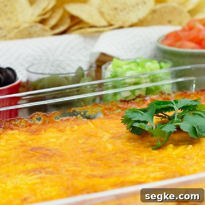 Clear glass casserole dish with beans topped with melted cheese and garnished with a sprig of cilantro. In the background are bowls of sliced black olives, jalapeno slices, diced green onions, diced tomatoes, and a basket of tortilla chips.