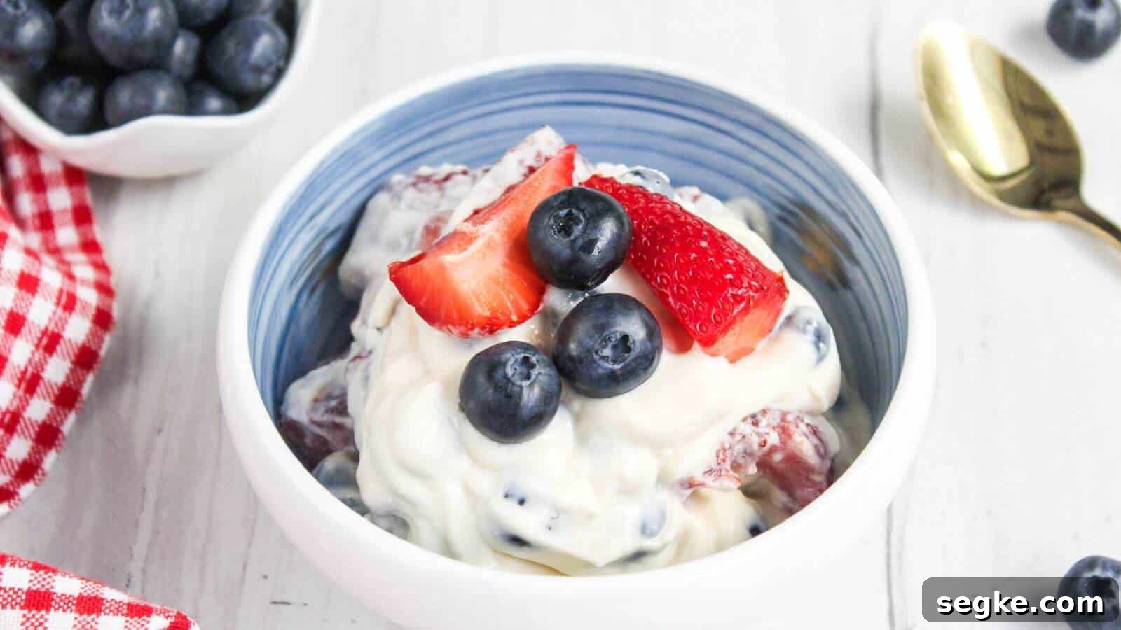 A bowl of creamy berry salad topped with fresh blueberries and sliced strawberries. A gold spoon is placed beside the bowl, and there is a small dish of blueberries in the background.