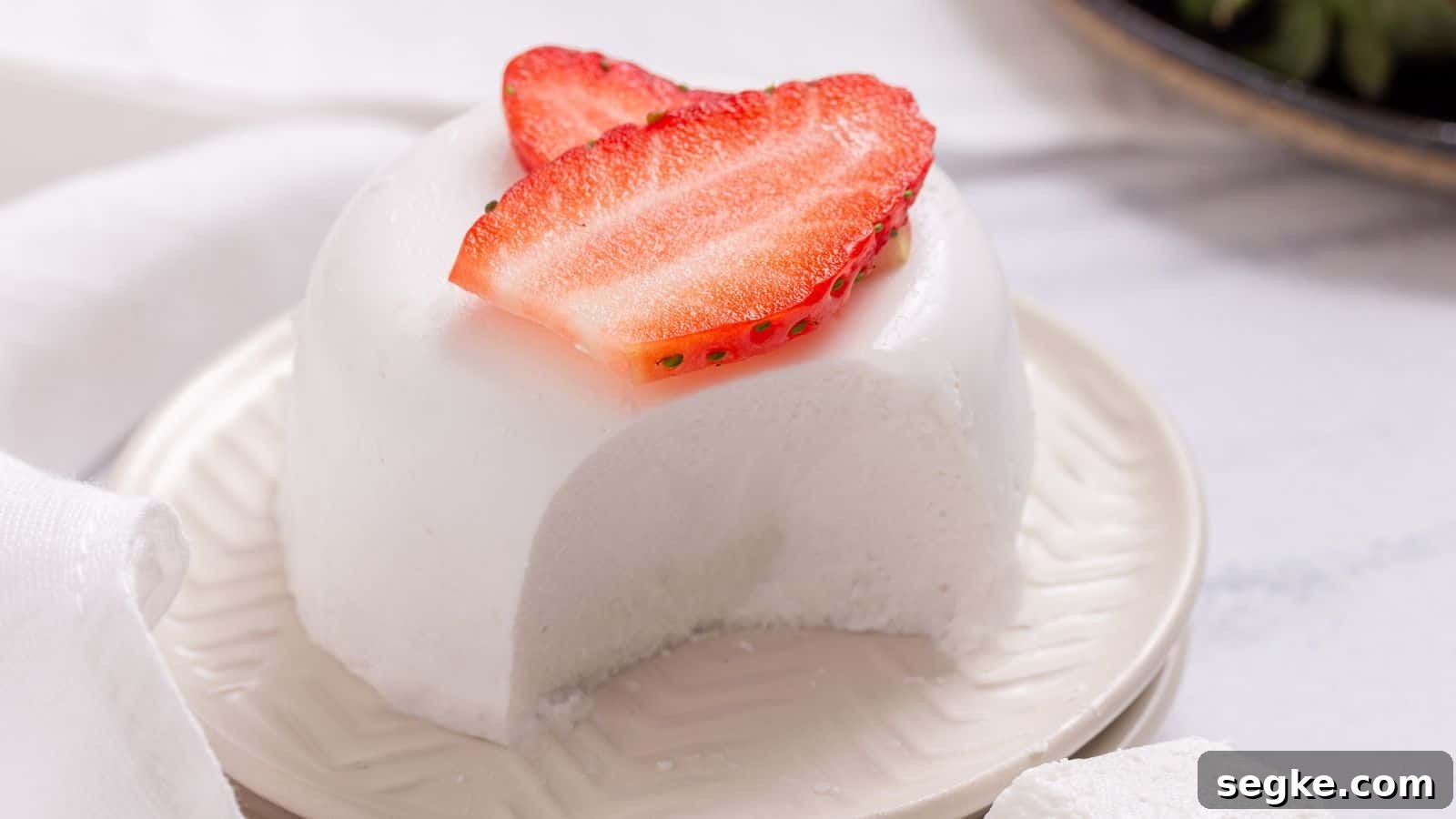 A slice of white, creamy panna cotta topped with a fresh strawberry slice on a white plate, with a textured napkin beside it.