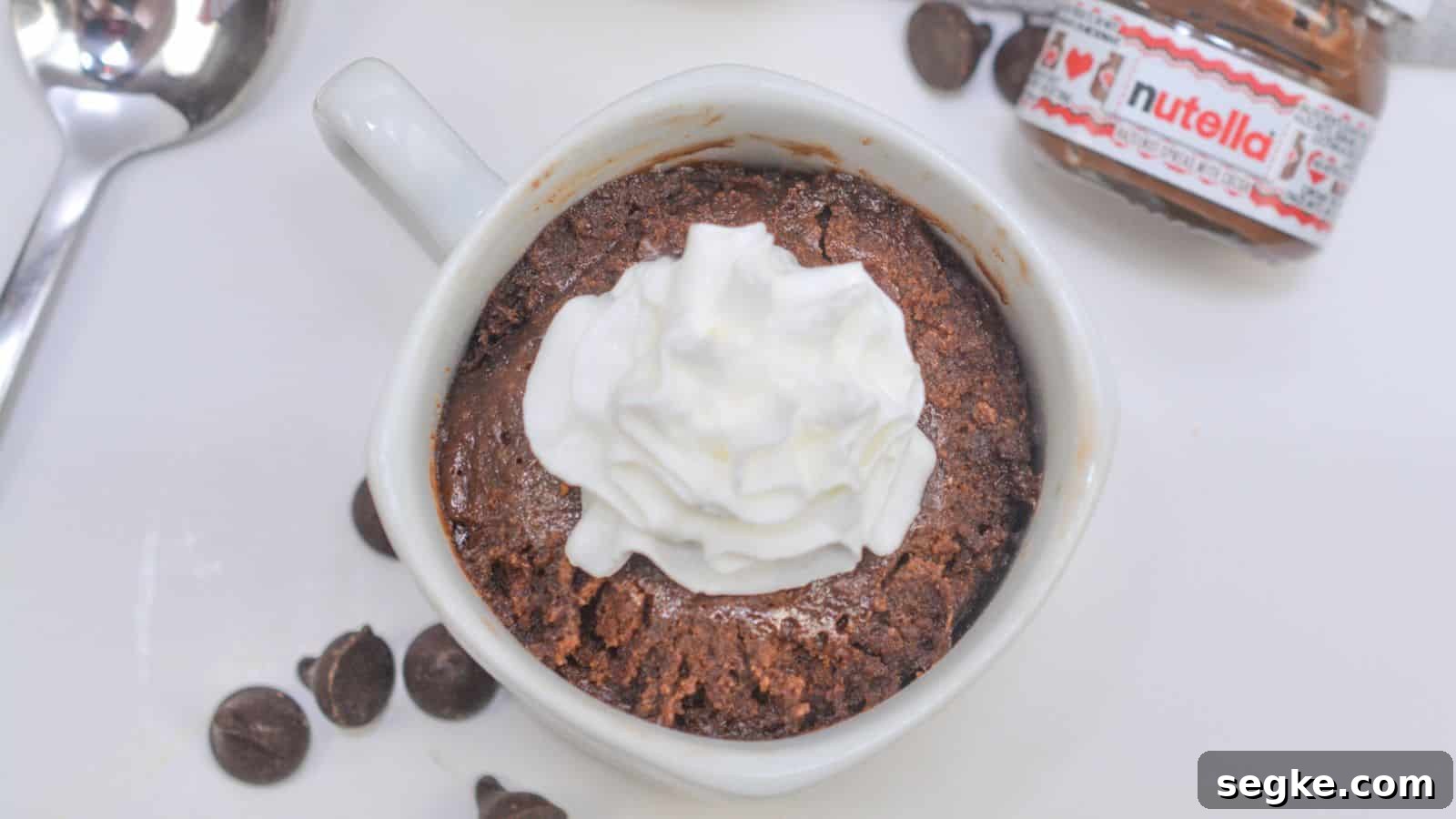 Homemade chocolate mug cake topped with whipped cream, served with a spoon and a small nutella jar on the side.