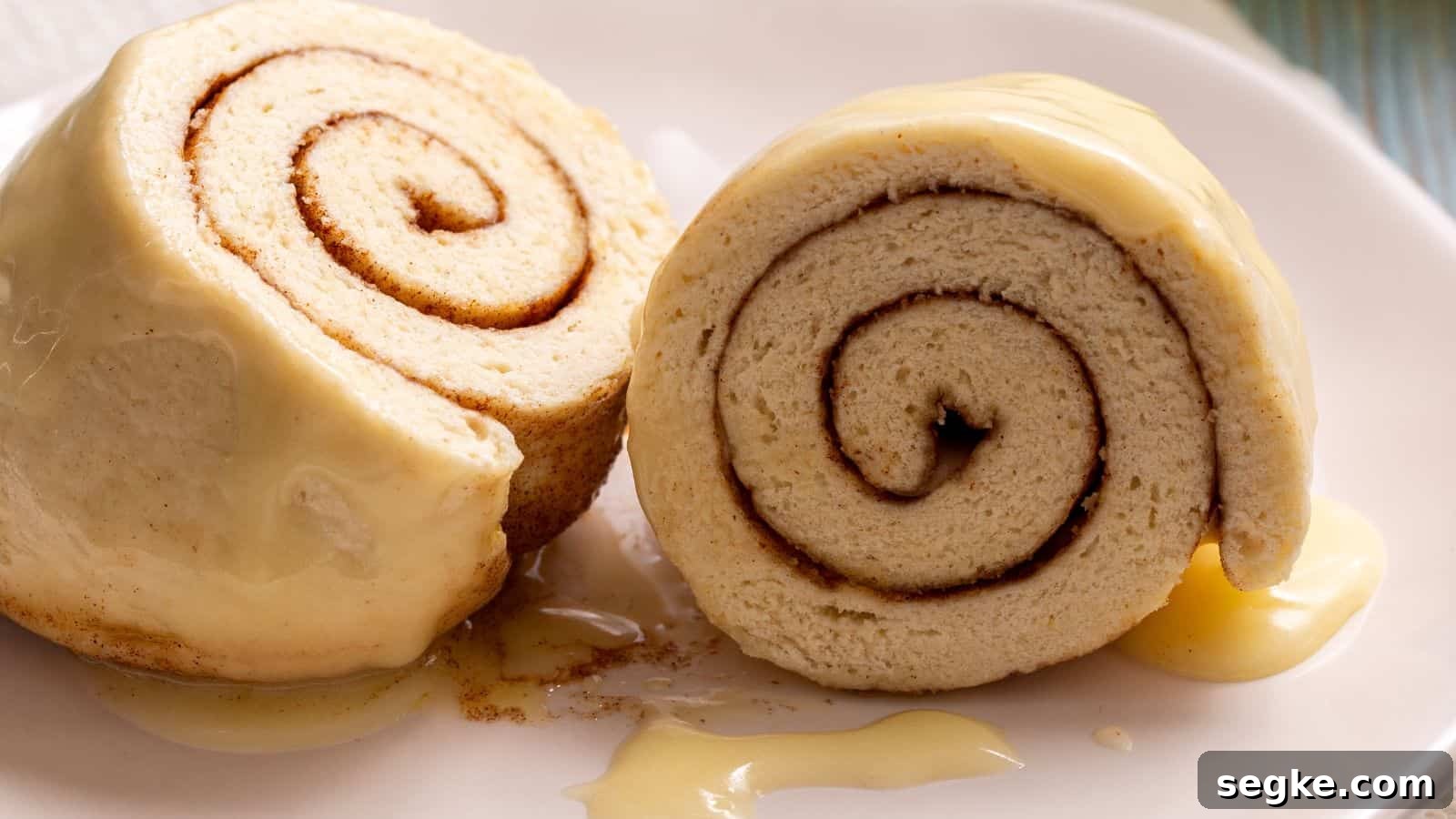 Two slices of cinnamon roll cake covered in creamy frosting, placed on a white plate with extra drizzle of sauce.