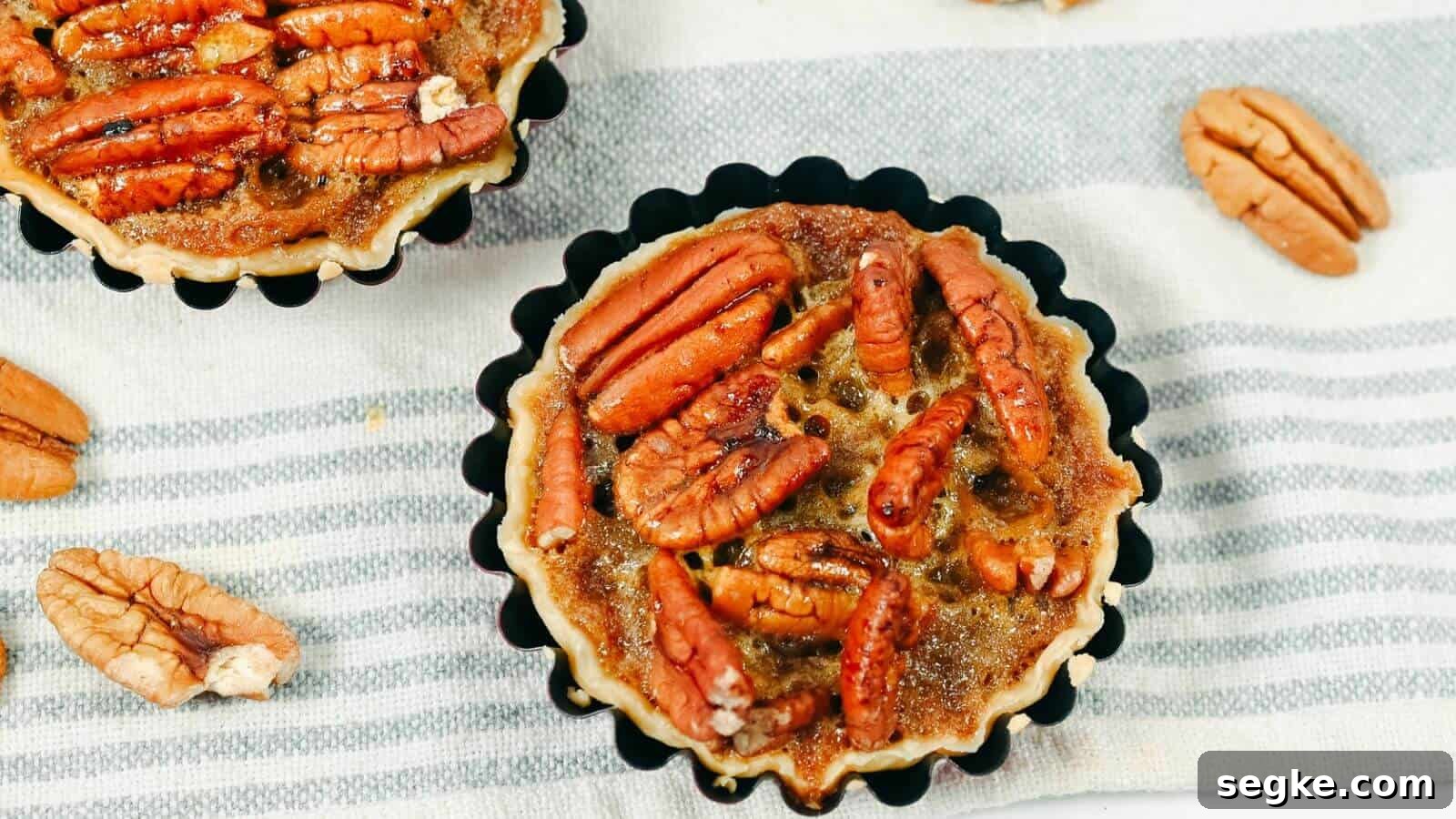 Two pecan pie tarts placed on a striped cloth with scattered pecans around them.