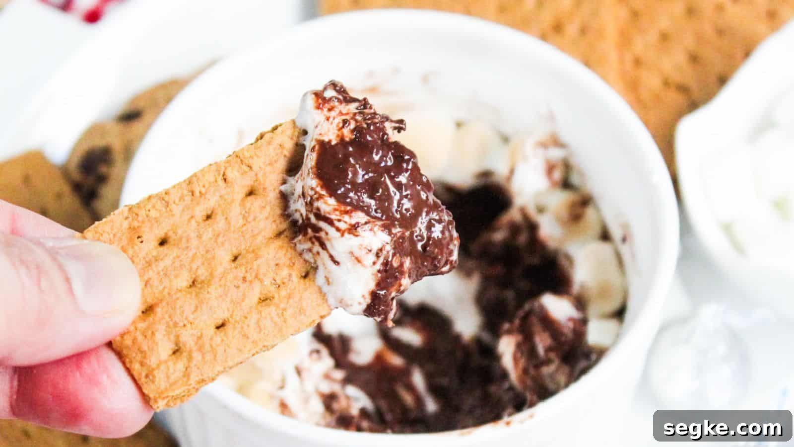 A hand holding a graham cracker dipped in s'mores dip with more crackers and dip in the background.