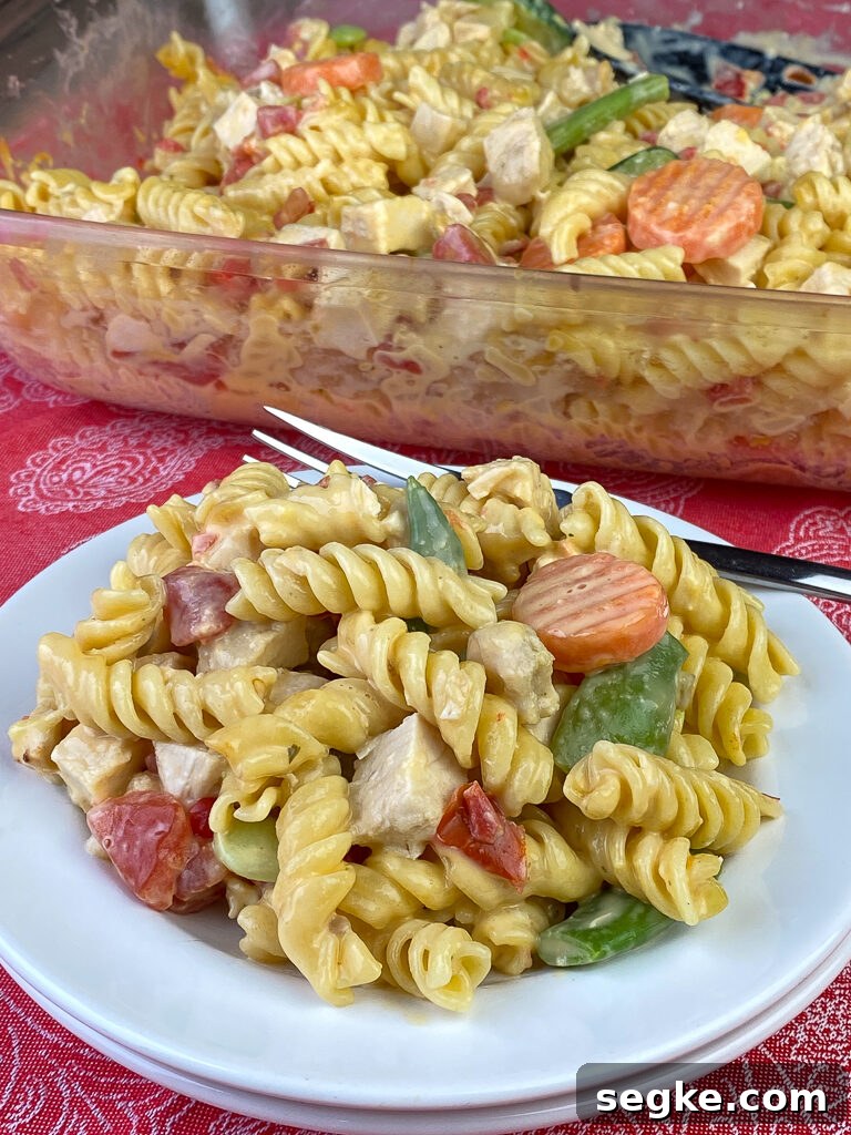 Plate full of cheesy Velveeta rotini pasta with tender pieces of chicken and veggies in a Cheesy Chicken Pasta Casserole with a fork and 9x13 dish full in the background.