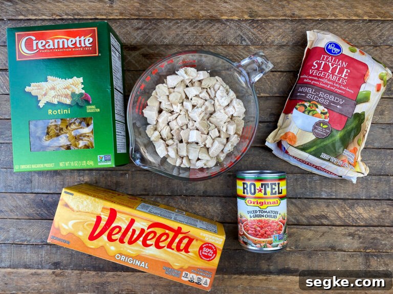 Ingredients for Velveeta chicken casserole are rotini, cubes of chicken, rotel, Velveeta and Italian style frozen veggies on a wooden counter top