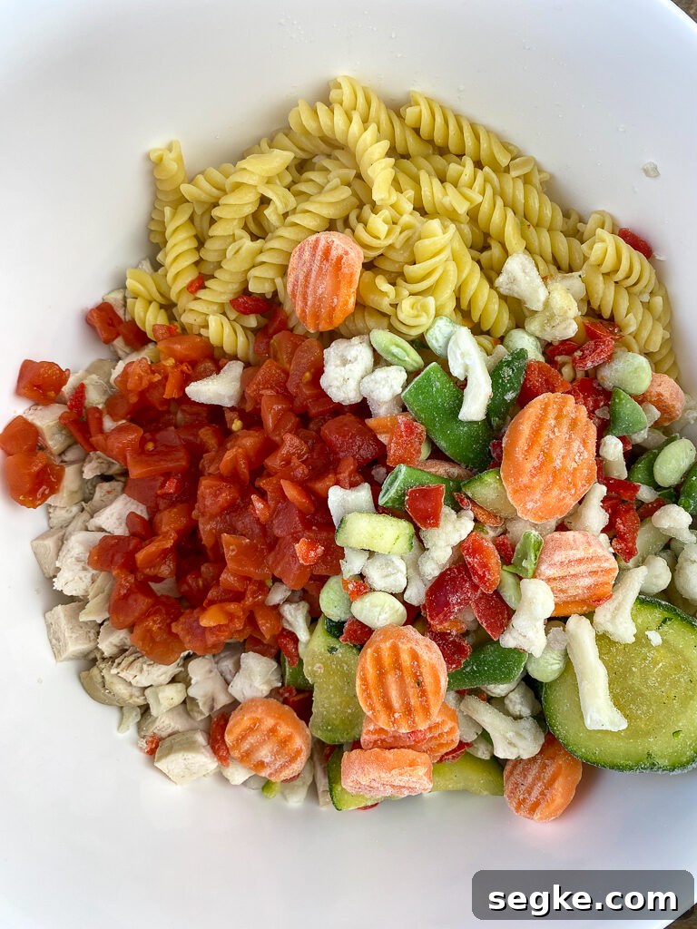Bag of frozen Italian style vegetables poured on top of pasta, cubes of chicken and can of rotel in a white mixing bowl
