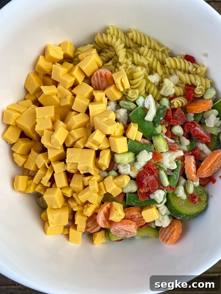 Cubes of Velveeta poured into a white mixing bowl with frozen mixed vegetables, cooked pasta, diced tomatoes and chicken