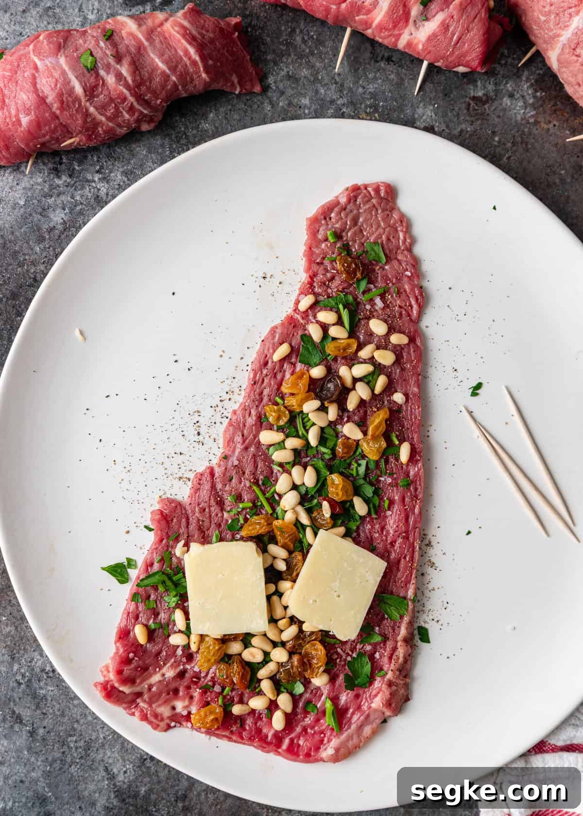 closeup overhead: flattened beef with seasoning, parsley, raisins, pine nuts, and cheese with toothpicks on white plate