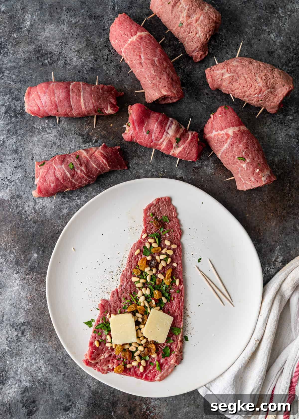 closeup overhead process shot: flattened beef with seasoning, parsley, raisins, pine nuts, and cheese with toothpicks on white plate with rolled braciole off to side