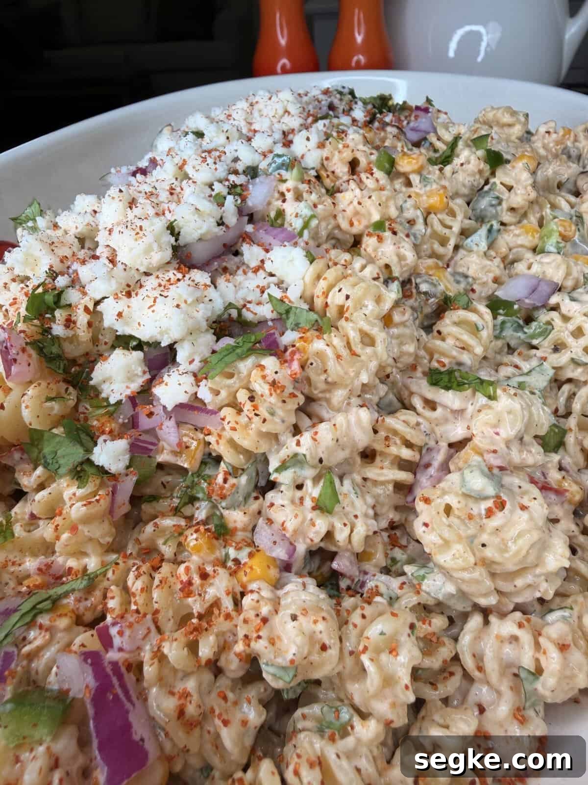 Mexican Street Corn Pasta Salad Recipe with Cotija and Lime 10 A close-up of a creamy pasta salad with rotini, chopped red onions, cilantro, crumbled cheese, corn, and a sprinkle of chili powder, served in a white dish with salt and pepper shakers in the background.
