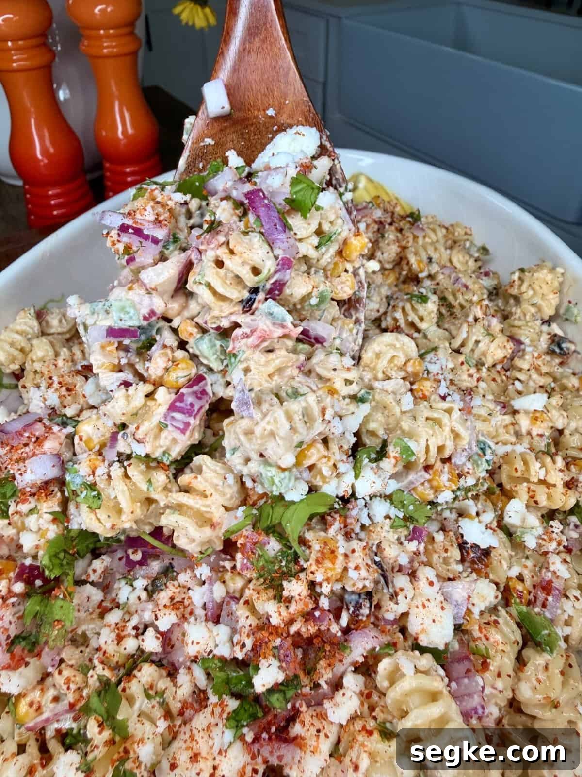 Mexican Street Corn Pasta Salad Recipe with Cotija and Lime 11 A wooden spoon scoops up creamy pasta salad with rotini noodles, red onion, corn, cilantro, cheese, and sprinkled spices from a white dish. Two pepper grinders are visible in the background.