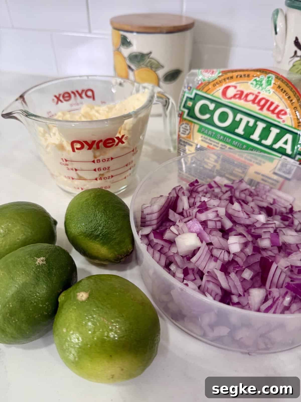 Mexican Street Corn Pasta Salad Recipe with Cotija and Lime 3 A kitchen counter with four limes, a container of chopped red onion, a measuring cup of mayonnaise, and a package of Cotija cheese. A decorative container with a lemon design is in the background.