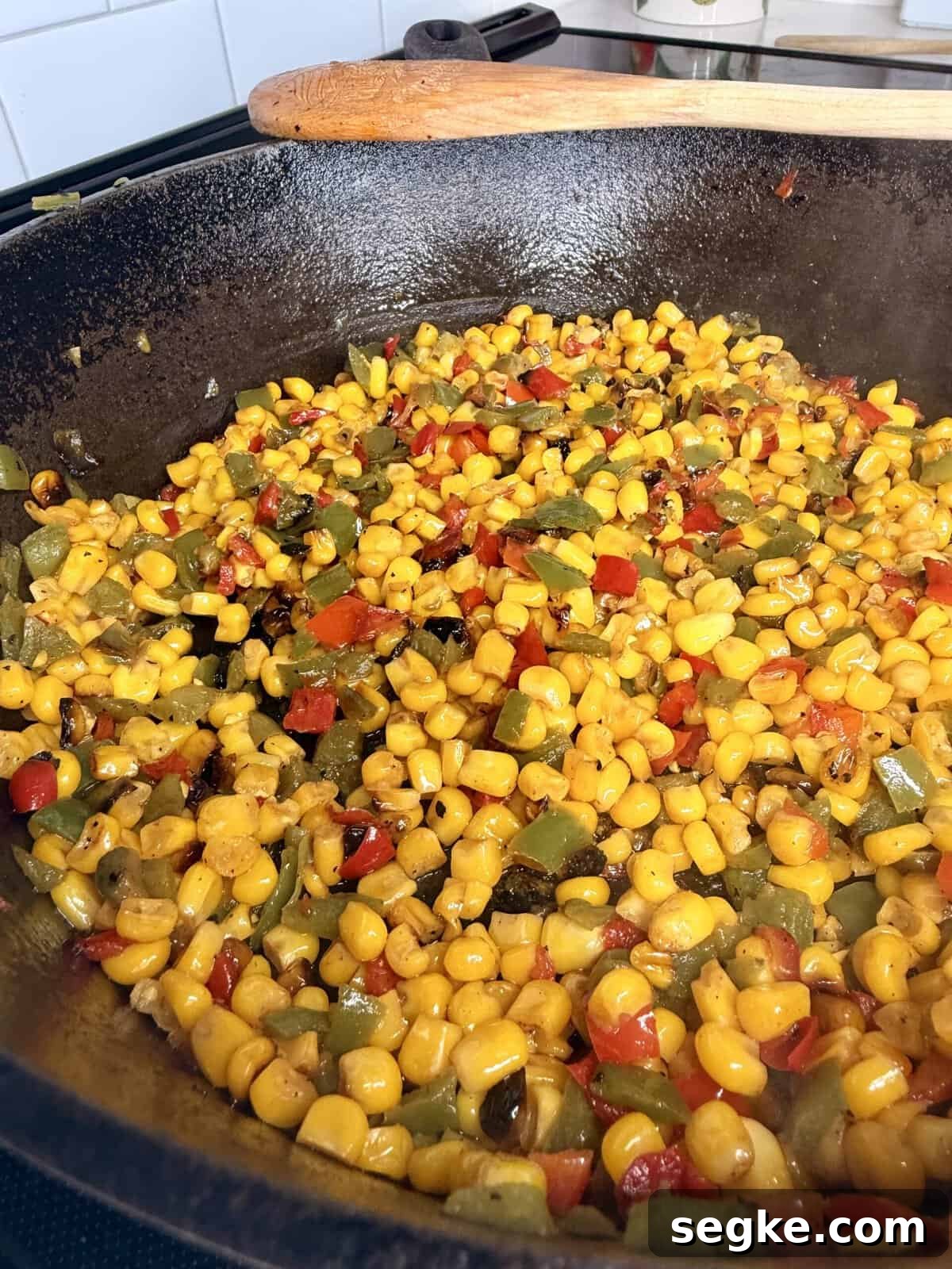 Mexican Street Corn Pasta Salad Recipe with Cotija and Lime 5 Colorful sautéed corn, red and green bell peppers, and onions cooking in a cast iron skillet on a stovetop, with a wooden spatula resting on the rim.