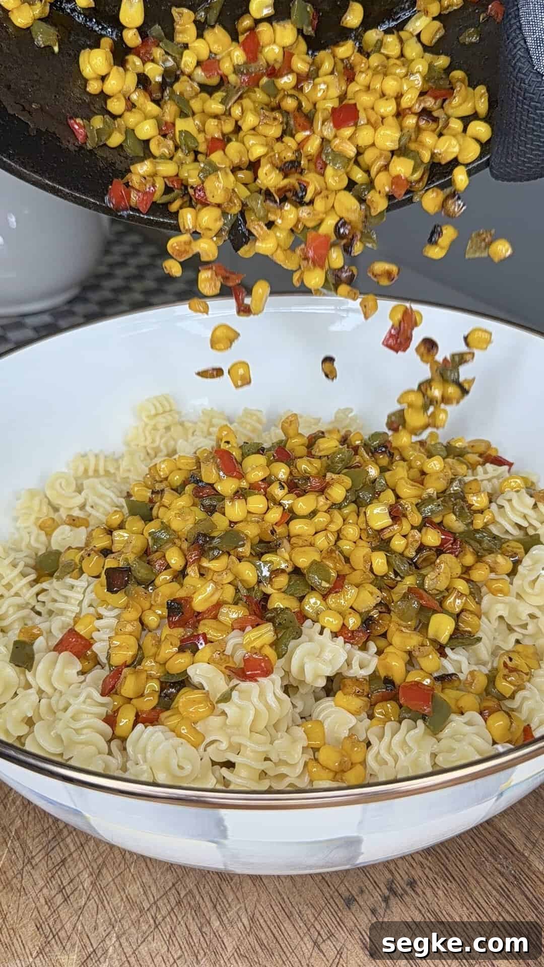 Mexican Street Corn Pasta Salad Recipe with Cotija and Lime 8 Charred corn and diced red and green peppers are being poured from a pan onto a bowl filled with cooked ruffle-edged pasta on a wooden surface.