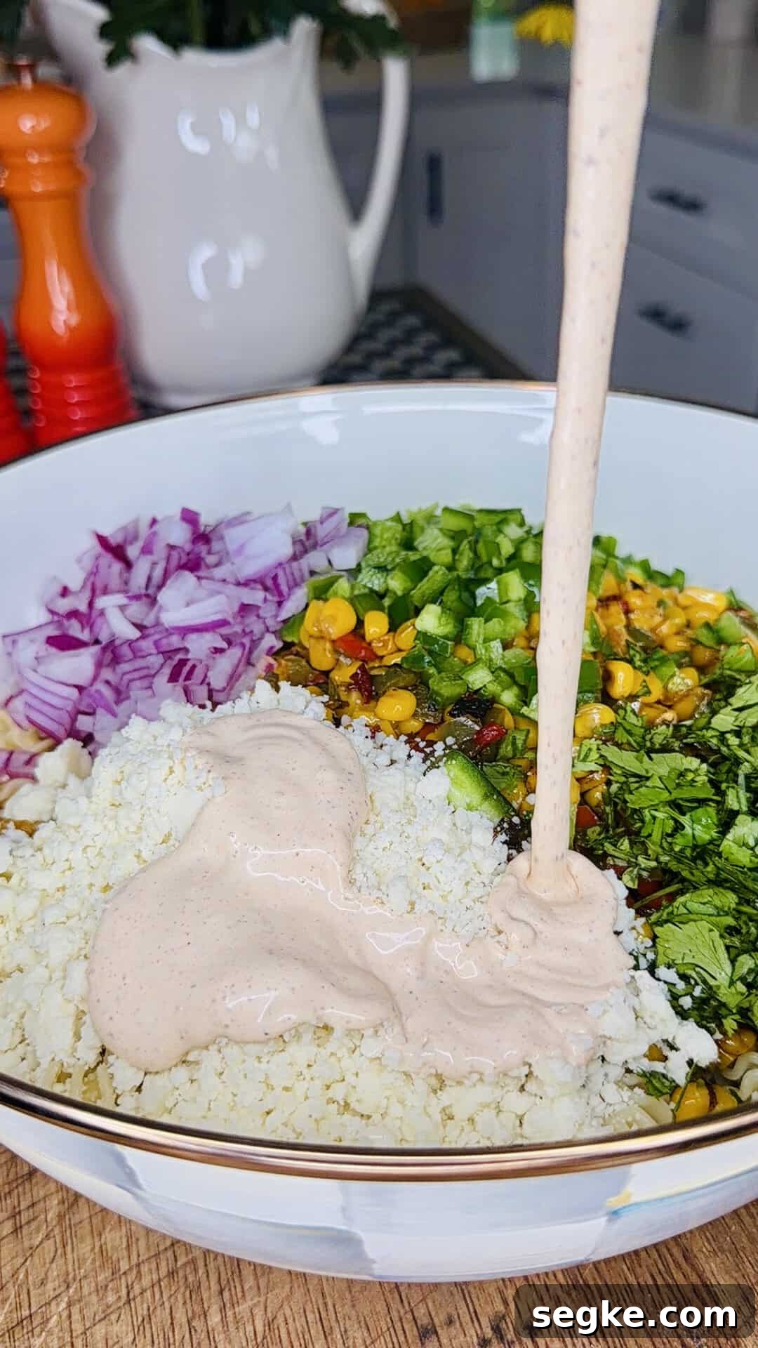 Mexican Street Corn Pasta Salad Recipe with Cotija and Lime 9 A bowl filled with chopped red onion, green peppers, corn, herbs, crumbled cheese, and a creamy sauce being poured on top, set on a wooden surface in a kitchen.