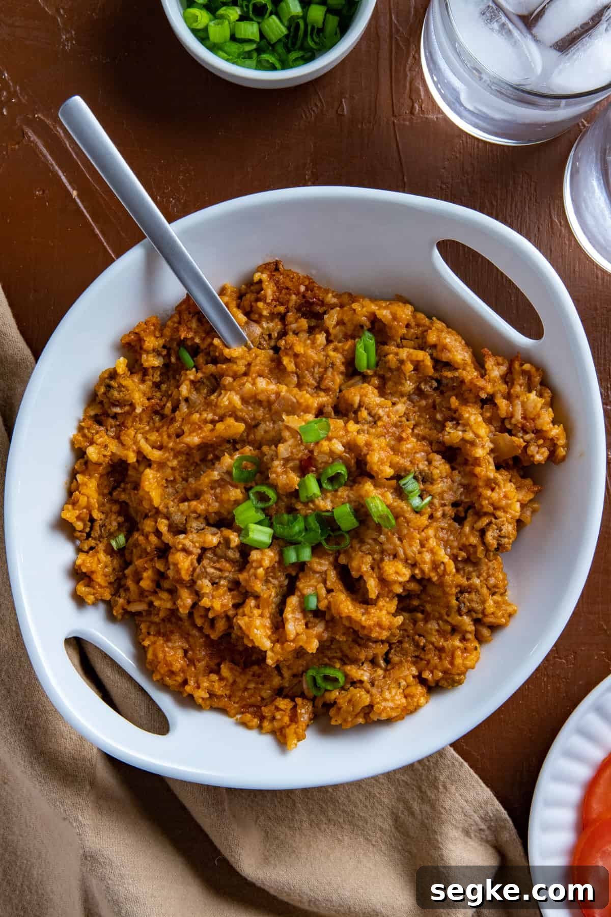 Cooked Spanish Rice in a white serving bowl with spoon inserted into the mixture. Sliced green onions on the side. Sliced tomatoes on the side.