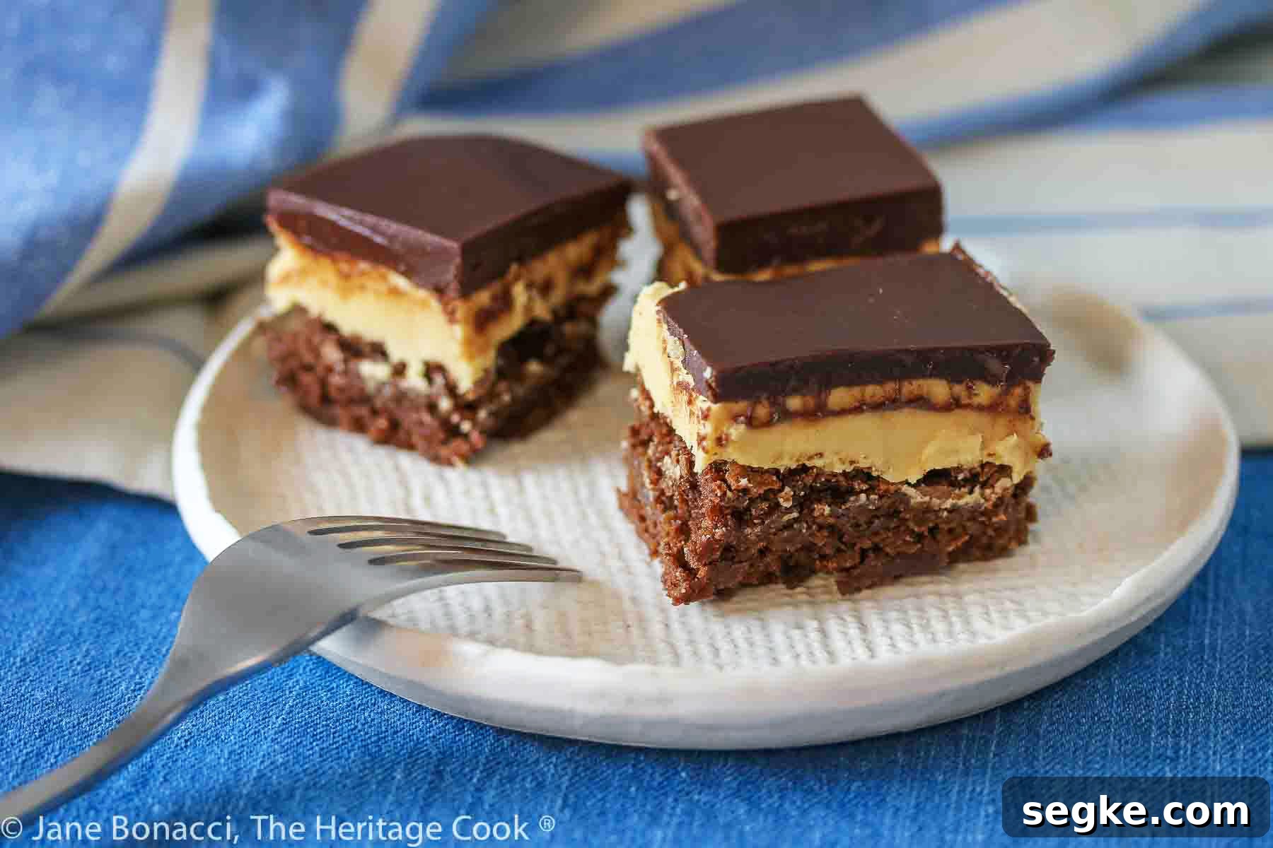 Three squares of layered Buckeye Peanut Butter Brownies on a white etched pottery plate set on a blue denim cloth with a blue striped cloth behind © 2024 Jane Bonacci, The Heritage Cook.