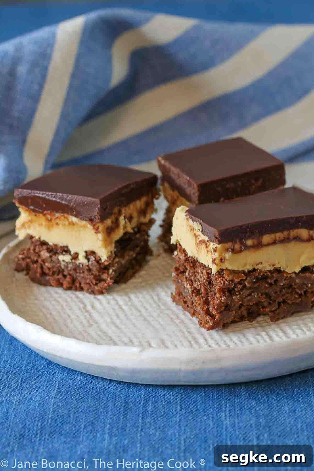 Three squares of layered Buckeye Peanut Butter Brownies on a white etched pottery plate set on a blue denim cloth with a blue striped cloth behind © 2024 Jane Bonacci, The Heritage Cook.