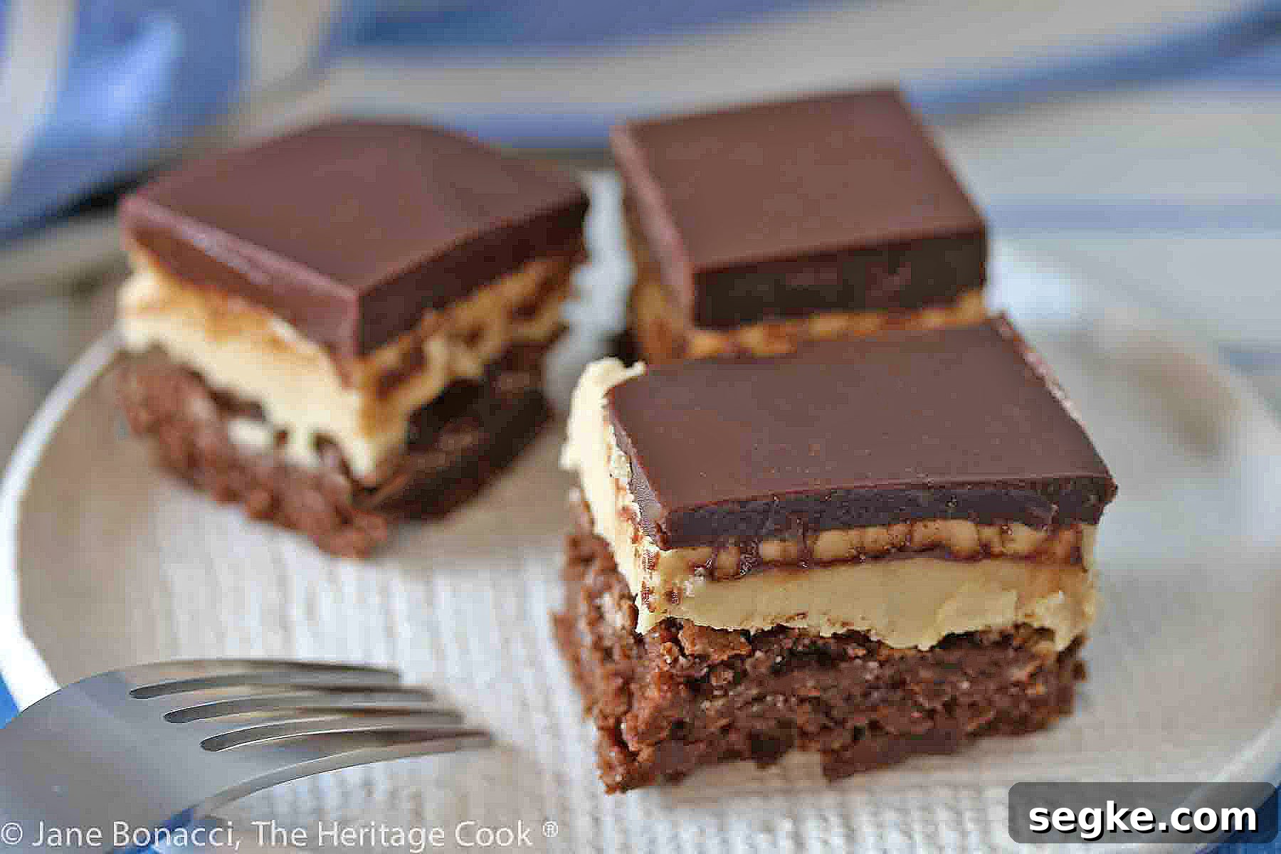 Three squares of layered Buckeye Peanut Butter Brownies on a white etched pottery plate set on a blue denim cloth with a blue striped cloth behind © 2024 Jane Bonacci, The Heritage Cook.