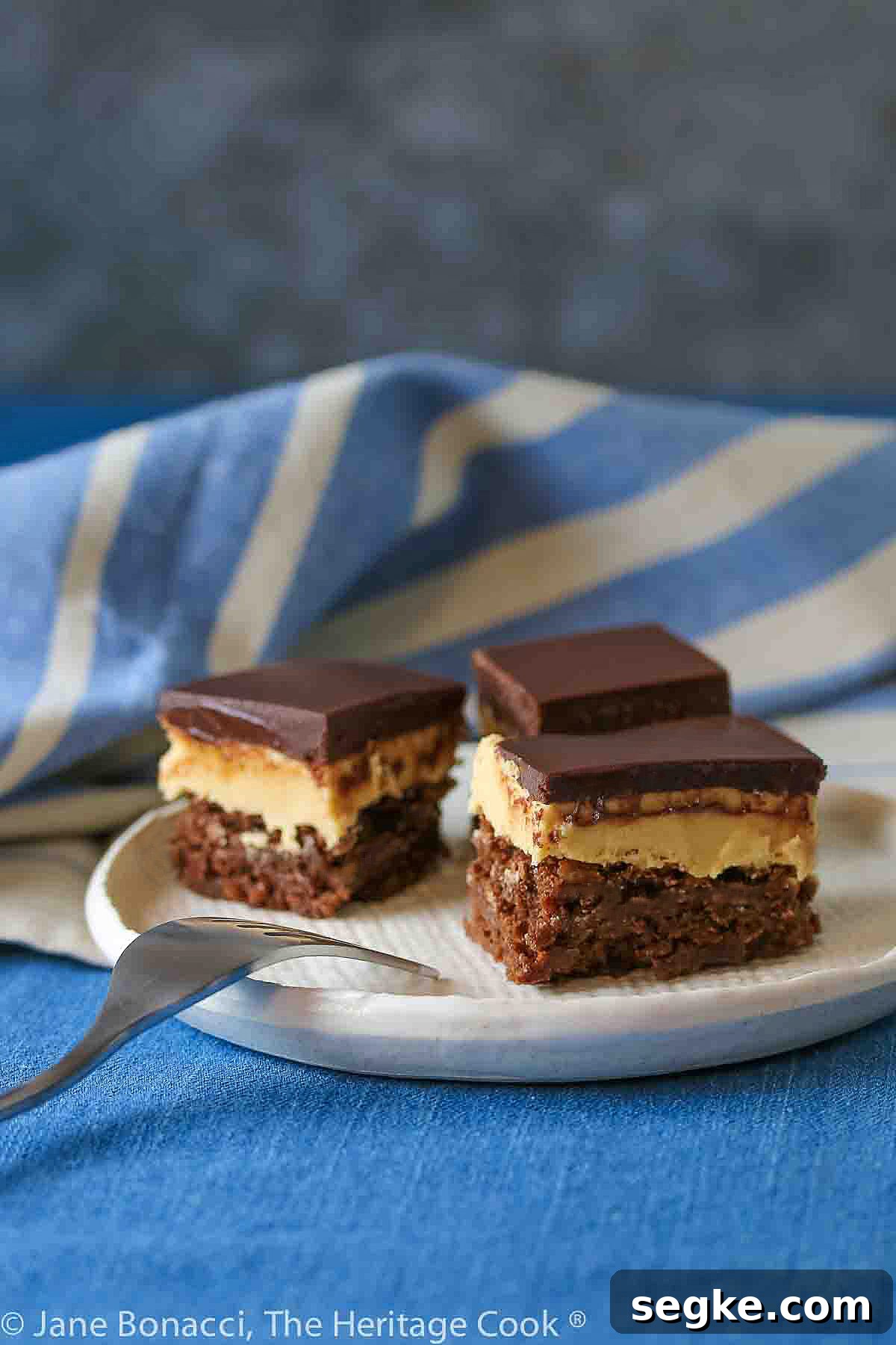 Three squares of layered Buckeye Peanut Butter Brownies on a white etched pottery plate set on a blue denim cloth with a blue striped cloth behind © 2024 Jane Bonacci, The Heritage Cook.