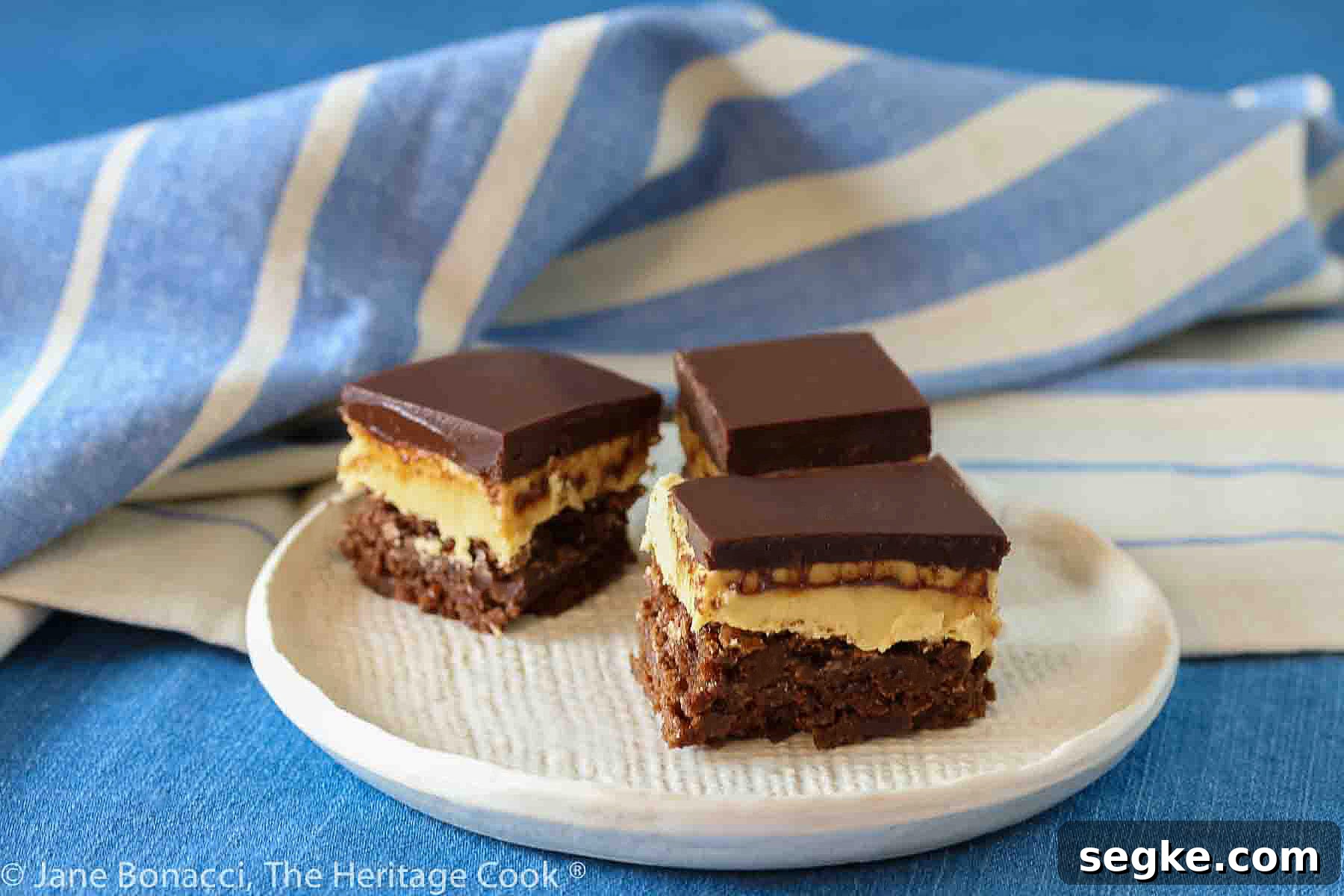 Three squares of layered Buckeye Peanut Butter Brownies on a white etched pottery plate set on a blue denim cloth with a blue striped cloth behind © 2024 Jane Bonacci, The Heritage Cook.