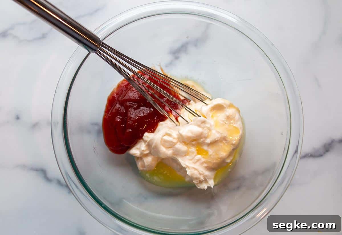 Glass bowl and whisk with mayo, melted butter, and ketchup. 