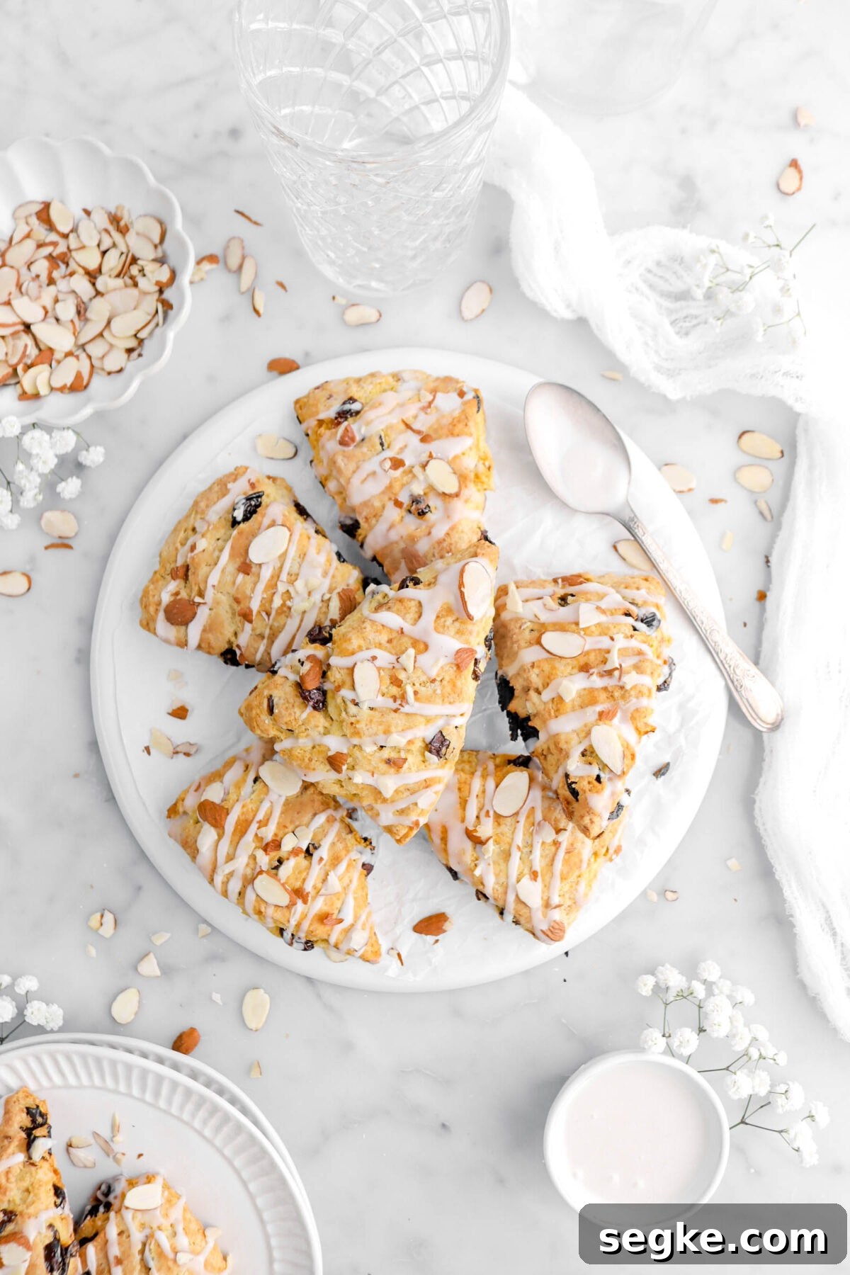 scones piled on upside down white plate with spoon beside, white flowers, almonds, and a cheese cloth around on marble surface.