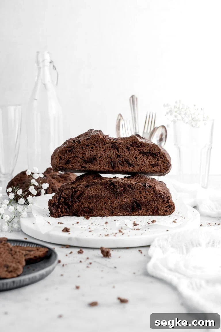 two halves of chocolate soda bread stacked on top of each other on marble surface with white flowers and crumbs around and another loaf of chocolate soda bread behind.